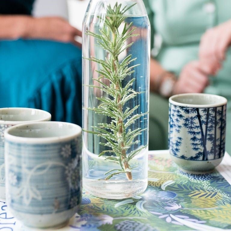 glass flask filled with water infused with sprig rosemary next to three ceramic cups