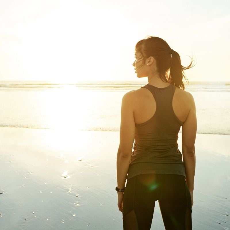 A woman in fitness gear looking at the sunset over the waves at a calm beach