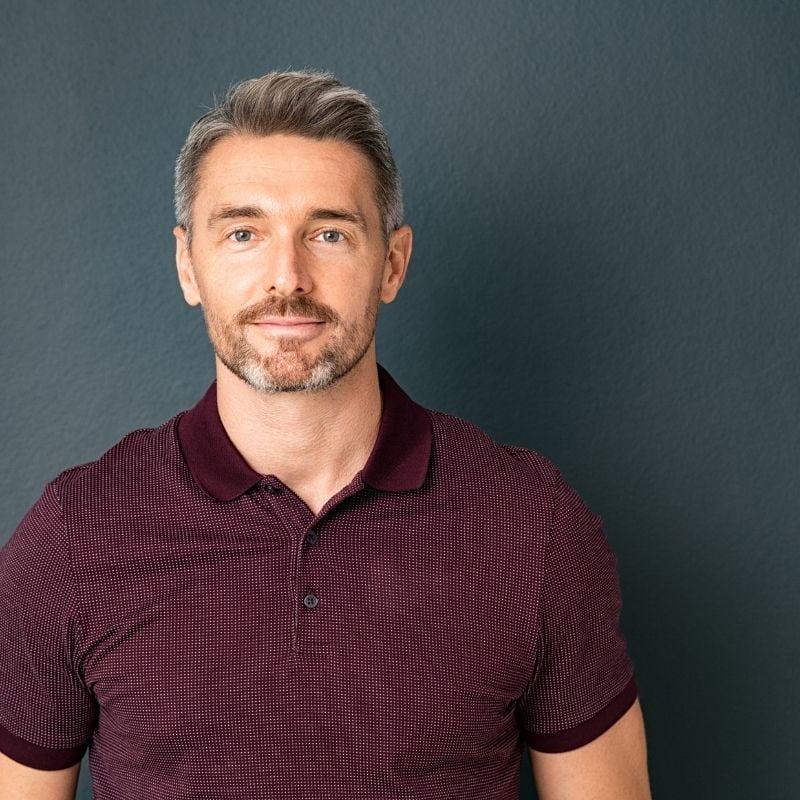 A fit middle aged man with greying hair wearing a polo top slightly smiling