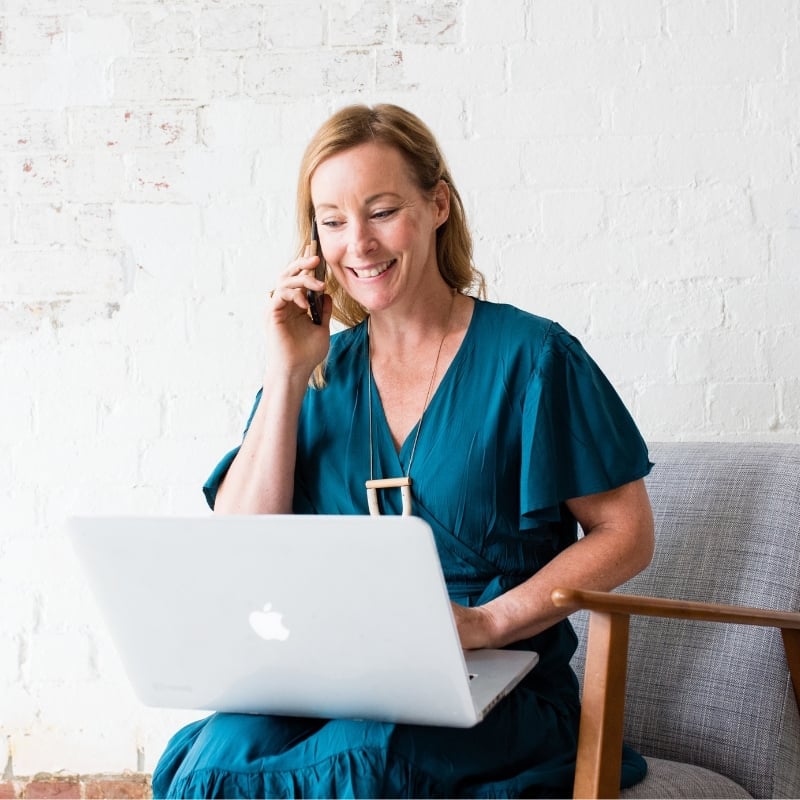 Health coach Bee Pennington wearing teal dress with phone against ear and looking at computer on her lap